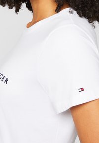 Zbliżenie na osobę w białej koszulce z krótkim rękawem, widocznym częściowym napisem "TOMMY HILFIGER" oraz małym wyszywanym logo na rękawie.