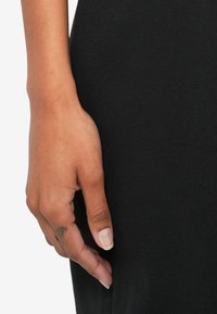 Melns apģērbs ar tuvplānu, kurā redzama roka, kas balstās uz tā. Audums šķiet gluds. Rokai ir gaiša nagu laka un redzama tetovējums.