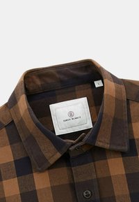 Chemise à carreaux marron et bleu marine foncé avec un col boutonné, une étiquette poitrine et une texture de tissu au fini doux. Taille L indiquée.