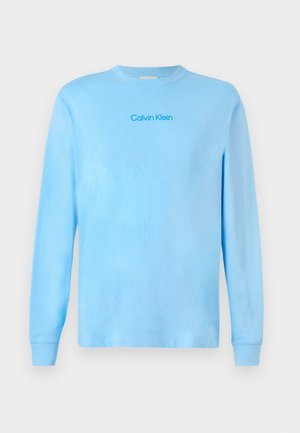 Hele sinine pikade varrukatega T-särk, millel on rinna keskosas "Calvin Klein" logo, esitatud lihtsal taustal.