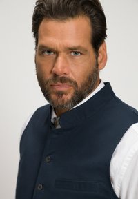 Chaleco sin mangas azul marino con cuello alto, hecho de tela texturada; presenta botones y un bolsillo en el frente, sobre una camisa blanca.