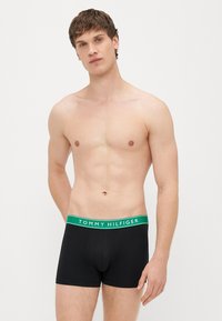 Boxers de algodão pretos com um cós verde e branco com o logótipo "TOMMY HILFIGER". Modelo em pé, sem camisa, mostrando a textura suave e o ajuste.