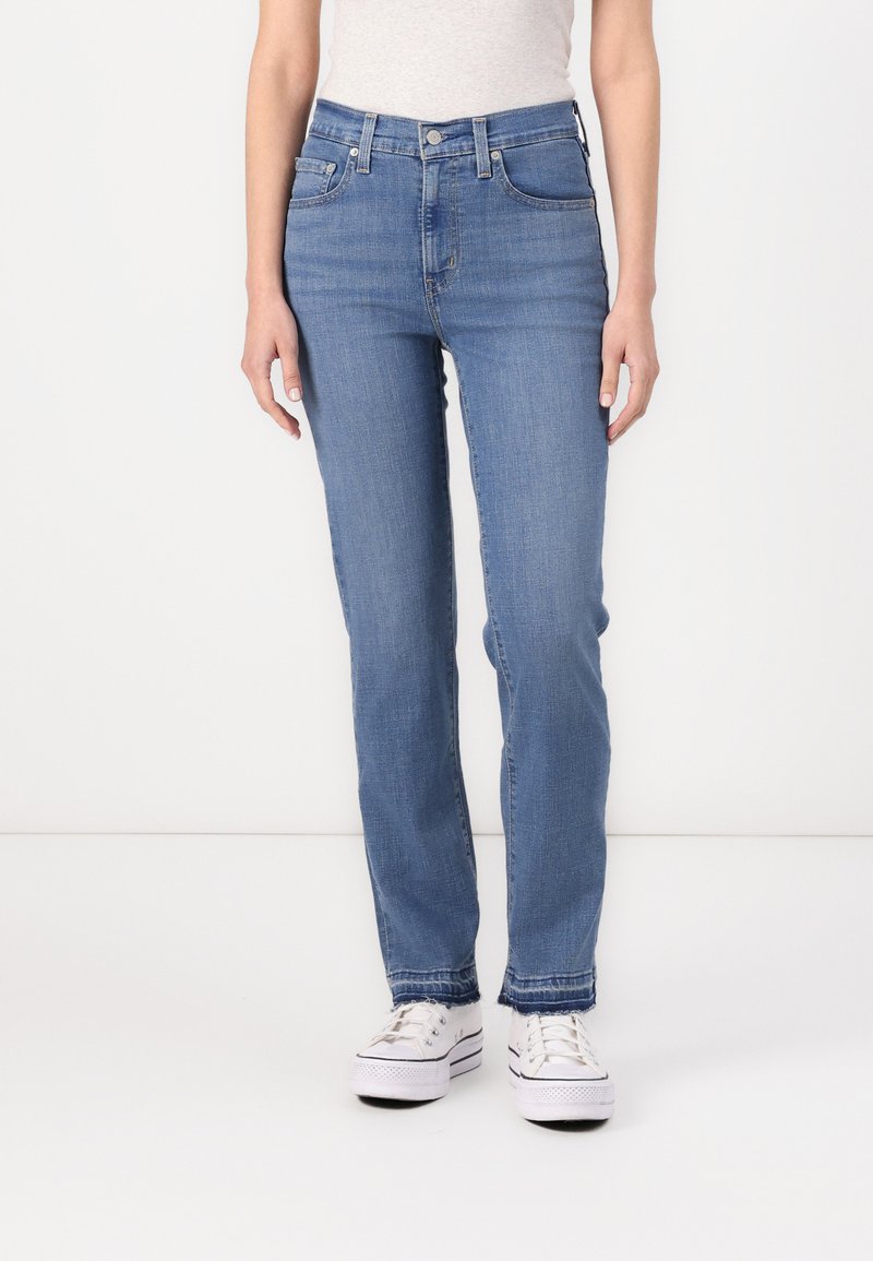 Eine Frau trägt hochgeschnittene blaue Jeans mit fransigen Säumen und weiße Plattform-Sneaker, während sie vor einem schlichten Hintergrund steht.