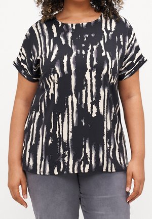 Femme portant un haut noir à manches courtes avec des rayures verticales blanches et abstraites, et un pantalon gris, debout les bras détendus le long du corps.