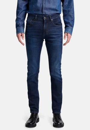 Dark blue denim jeans with a fitted design, five pockets, and a button closure. Paired with black lace-up shoes.