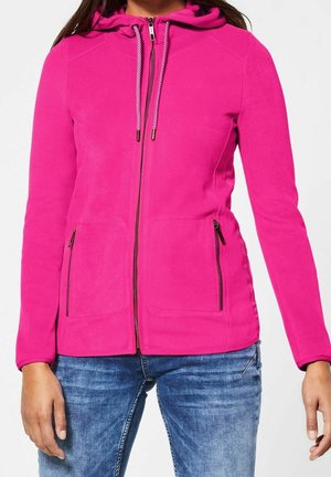 Woman wearing a bright pink zip-up hoodie with drawstrings and zippered side pockets, paired with blue jeans.