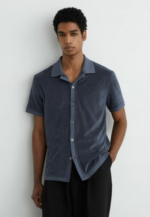 Jeune homme aux cheveux courts et bouclés, debout contre un mur blanc, portant une chemise gris foncé à manches courtes boutonnée et un pantalon noir.