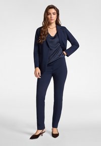 Traje azul marino con blazer a medida y pantalones slim-fit, combinado con una camisola de satén y tacones slingback marrones. Acentuado con una cadena dorada.