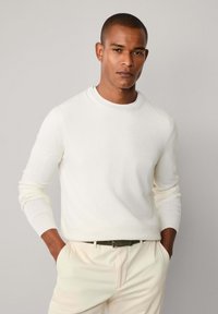 Muškarac s kratkom kosom odjeven u bijeli pulover s okruglim izrezom i svijetlo bež hlačama, stoji s rukama u džepovima na svijetlosivoj pozadini.