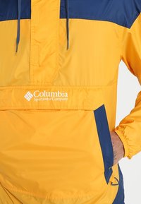 Sudadera amarilla y marina con un bolsillo frontal, capucha ajustable con cordón y logotipo en el pecho, fabricada en material ligero.