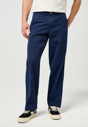 Uomo che indossa pantaloni dritti blu navy e sneakers nere con suole bianche, in piedi contro uno sfondo chiaro e uniforme.