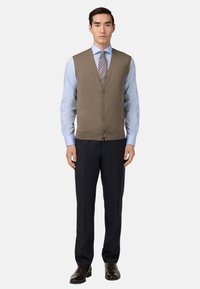 Hnedý pletený sveter cez svetlomodrú košeľu, skombinovaný s tmavými prúžkovanými nohavicami a lesklými čiernymi formálnymi topánkami. Zapínanie vpredu a strih na mieru.