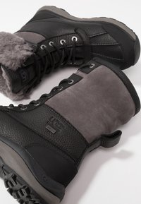 Para czarnych i szarych wodoodpornych zimowych butów UGG z sznurowaniem, owczą podszewką i gumowymi podeszwami, na białym tle.