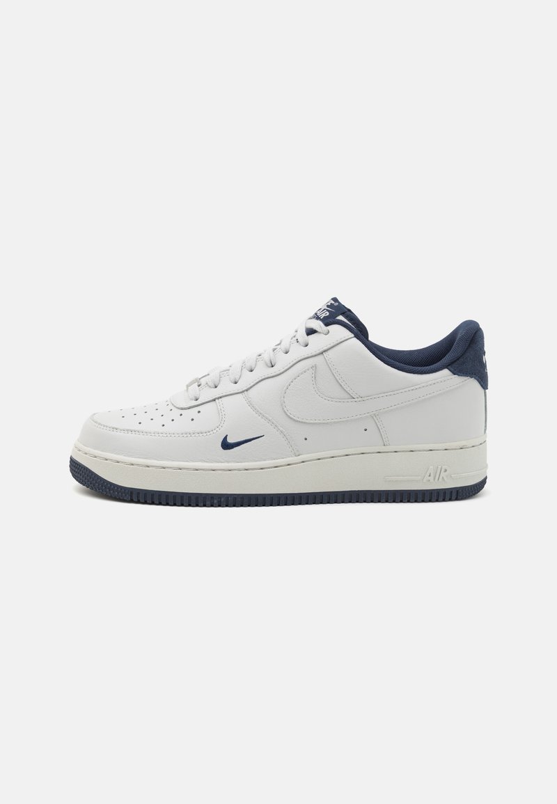 Fehér bőr Nike Air Force 1 sportcipők sötétkék kiegészítőkkel, perforált orral, lapos fűzőkkel és texturált gumitalppal.