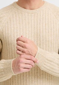 Suéter de punto acanalado beige con cuello redondo. La tela texturada presenta patrones verticales y los puños ajustados acentúan las mangas.