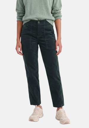 Personne portant un pantalon droit vert foncé avec des poches plaquées à l'avant, un pull vert clair et des baskets beiges, debout sur fond blanc.