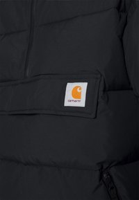 Svart dunjacksmaterial med quiltad mönster. Har en vit lapp med en orange logotyp på vänster bröst. Dragkedjeficka på sidan.