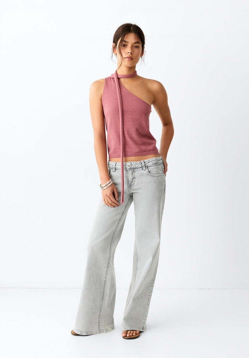 Femme portant un top rose à une épaule avec un long ruban et un jean ample gris clair, debout avec une main dans la poche et des sandales.