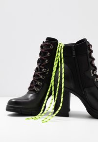 Bottes basses noires à finition brillante, avec des lacets rouges et noirs, un talon bloc haut et des lacets décoratifs jaunes fluo à l'arrière.