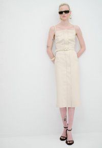 Robe midi ajustée beige avec des bretelles spaghetti, taille ceinturée, fente avant et poches latérales. Portée avec des talons noirs et des lunettes de soleil.
