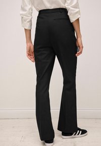 Pantalons noirs évasés avec une texture lisse, présentant une poche arrière unique et associés à des baskets noires, le tout sur un fond neutre.