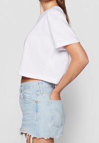 Femme portant un t-shirt court blanc et une jupe en jean bleu clair usée avec un ourlet effiloché, mains dans les poches de la jupe.