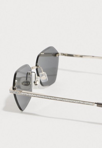 Lunettes de soleil hexagonales avec des verres gris foncé et une monture en métal argenté. Accents décoratifs en argent sur les branches et les coussinets de nez.