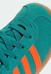 Teal mockascu sneaker med orange zigzag-ränder, platta teal-snören och en gummisula med texturerat grepp. Närbild av överdelens design.