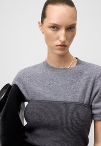 Jeune femme avec les cheveux plaqués en arrière, portant un haut en maille gris bicolore et un collier de perles, tenant un grand sac en cuir noir.