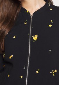 Černá zipová bunda s vysokým límcem, zdobená drobnými žlutými květinovými vzory rozptýlenými po celém povrchu. Měkká tkanina s hladkým povrchem.