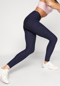 Mørkeblå leggings med teksturfinish, høy midje og sømdetaljer, kombinert med hvite joggesko med lilla detaljer.