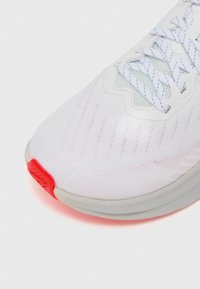 Chaussure de course avec une tige en mesh blanc, des lacets en textile et un accent de semelle en caoutchouc rouge. Présente un design léger et une surface texturée.