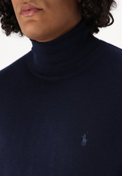 Tumši zils turtlenecks džemperis mīkstā adījumā, ar mazu izšūtu logotipu uz krūtīm un pieguļošu dizainu.