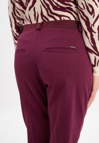 Burgundy trousers with a smooth texture, featuring a logoed waistband and two back pockets. The design is tailored for a fitted look.