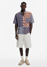 Short-sleeve shirt features a patchwork design with geometric patterns in blue, orange, and stripes, paired with cream shorts and beige shoes.