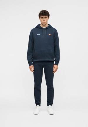 Mladý muž stojaci, oblečený v tmavomodrej mikine s kapucňou a logami, k tomu ladivé tepláky a biele tenisky, na jednoduchom bielom pozadí.