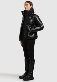 Veste matelassée noire avec col montant, finition brillante et poches zippées. Portée avec un legging noir et des bottes hautes jusqu'au genou.