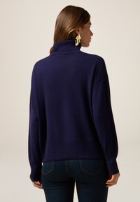 Dunkelblauer Pullover mit hohem Kragen, weicher Textur und lockerer Passform. Das Modell trägt enganliegende Jeans und zeigt einen klassischen, lässigen Look.