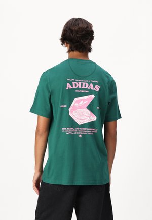 Mężczyzna w zielonej koszulce Adidas z różowym nadrukiem pizzy i napisem na plecach, w czarnych szortach, odwrócony tyłem do kamery.