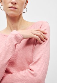 Dlhý rukáv top v mäkkej svetloružovej látke, predná časť má uvoľnený výstrih a mierne textúrovaný povrch. Rukávy sa zužujú pri zápästiach.