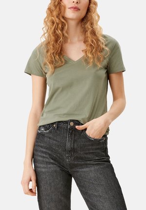 Woman with long curly hair wearing an olive green short-sleeve V-neck T-shirt and high-waisted faded black jeans, hand in pocket.
