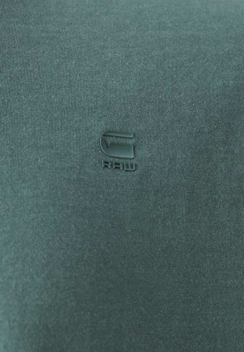 Grünes Baumwoll-Crewneck-T-Shirt mit einem kleinen, geprägten Logo in der Mitte. Der Stoff hat eine weiche, glatte Textur mit einem leichten Glanz.