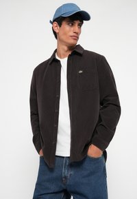 Chemise en velours côtelé marron avec un devant à boutons, une poche poitrine et le logo Lacoste sur le côté gauche, associée à un t-shirt blanc et un jean en denim bleu.