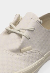 Chaussure Vans de couleur blanc cassé avec un motif à damier texturé et des lacets assortis, mettant en avant le côté supérieur et une partie de la semelle de la sneaker.