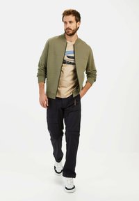 Olijfgroene bomberjack met gestructureerde panelen, beige t-shirt met blauwe grafische print, marineblauwe cargobroek en witte sneakers met zwarte accenten.