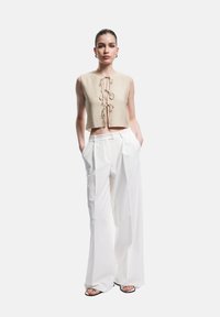Top cropped beige avec un nœud à l'avant, associé à un pantalon large blanc. Texture lisse et coupe décontractée. Sandales noires à brides.
