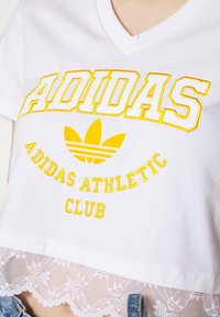 Vit bomull t-shirt med broderad "ADIDAS" logotyp i gult och texten "ADIDAS ATHLETIC CLUB", med en scalloped spetsfåll.
