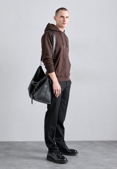 Hanorac brown cu buzunar frontal, geantă de umăr neagră, pantaloni negri și pantofi negri. Modelul stă pe o suprafață gri, pe un fundal simplu.