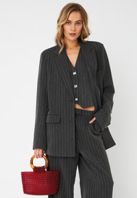 Completo grigio a righe con giacca doppiopetto, top corto e pantaloni a gamba larga. Modella che tiene in mano una piccola borsa di perline rosse con manico rotondo.