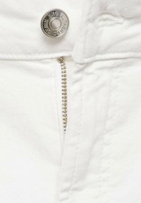 Detail bielej denimu zošitého zblízka, so strieborným gombíkom a kovovým zipsom so zlatými zubami. Hladký povrch s jemným prešitím.